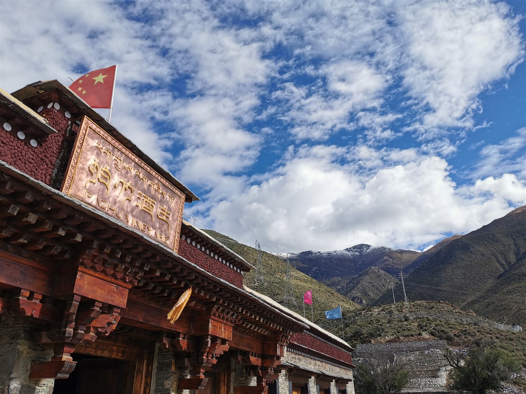 The estate gate: a copper plaque lettered in Tibetan, Chinese and English above carved timber eaves, prayer flags and snow mountains beyond